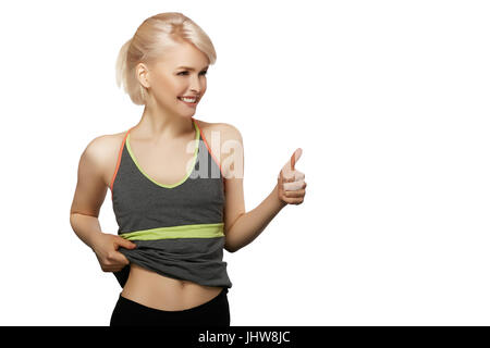 Slim ragazza in abbigliamento sportivo mostra il suo ventre e il pollice in alto isolato su sfondo bianco Foto Stock