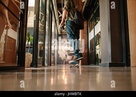 Basso angolo vista posteriore colpo di uomo informale skateboarding in ufficio. Giovane imprenditore pattinaggio attraverso il suo ufficio di avvio. Foto Stock