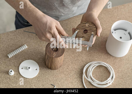 Le mani di un uomo con una pinza che è la misurazione di una dimensione di un round billetta in legno in officina. Sotto le sue mani ci sono di metallo e legno di forma cilindrica Foto Stock