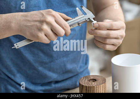 Il processo di misurazione in officina. L uomo è utilizzando una pinza sul metallo bianco dettaglio. Sotto le sue mani ci sono in legno e metallo billette cilindrico. C Foto Stock