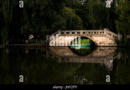 Arcuata di ponte di pietra e la sua riflessione sopra il lago verde in Kunming Cina circondato da alberi Foto Stock