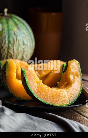 Mature succose fette di melone arancione, piastra scuro, coltello, legno tavolo da cucina, arredamento rustico stile minimalista Foto Stock