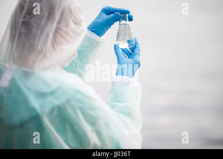 Biologo foto dalla parte posteriore e in tuta protettiva con lampadina con acqua in mani sulla banca del fiume Foto Stock