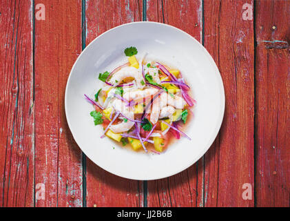 Aggiornamento piatto di pesce marinato nel succo di agrumi. Gamberi e Mango Ceviche su rosso tavolo in legno. La dieta e il cibo sano concetto Foto Stock