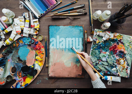 Pittore workplace top view photo. Artista pennelli e colorati di colori e pastelli assortita sulla scrivania. Foto Stock