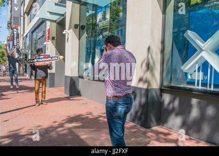 Un maschio Caucasion fotografo spara un afro-americano di uomo il cui compito è quello di portare un cartello per il Camp Wannaplaya intorno al Market Street a San Fran Foto Stock