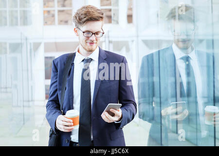 Imprenditore utilizza lo smartphone e di bere il caffè nei moderni interni luminosi di ufficio corridoio di vetro Foto Stock