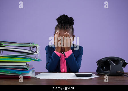 Imprenditrice che copre gli occhi mentre è seduto alla scrivania contro il muro viola in office Foto Stock