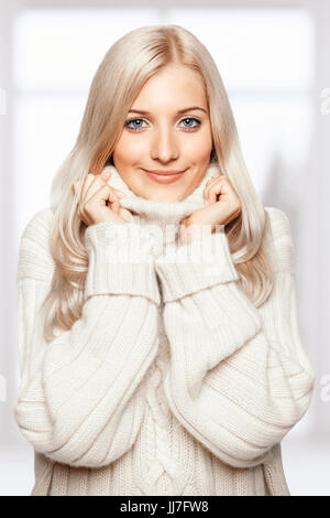 Bionda giovane bella donna vestita di bianco grande maglione cashmere su sfondo finestra Foto Stock