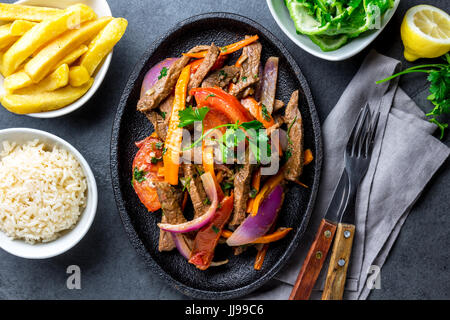 Piatto peruviano lomo saltado - filetto di manzo con viola cipolla, peperoncino giallo, pomodori servita su piastra nera con patatine fritte e riso. Vista superiore Foto Stock