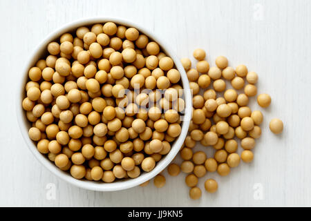Asciugare i semi di soia in ceramica bianca recipiente isolato sul dipinto di bianco legno proveniente da sopra. Eventuali fuoriuscite di fagioli. Foto Stock