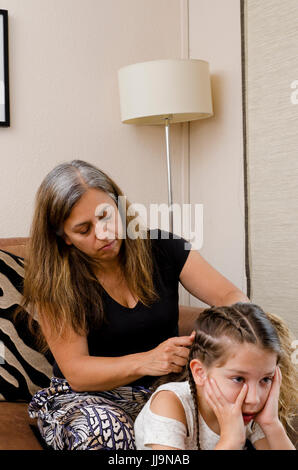 Madre e figlia e lo stile di vita della famiglia genitorialità mostrante una stanca mamma styling la sua giovane figlia di capelli in trecce a casa. Foto Stock