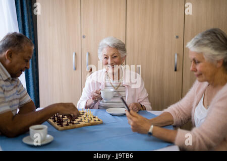 Donna sorridente con il caffè seduti con gli amici a giocare a scacchi e utilizzando la tecnologia a tavola in casa di cura Foto Stock