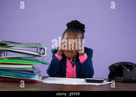 Stanco imprenditrice che copre gli occhi mentre è seduto alla scrivania contro il muro viola in ufficio Foto Stock