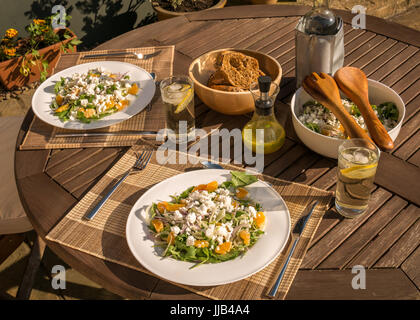 Insalata estiva servita fuori in giardino patio con tavolo clementine, rucola, finocchio, cicoria, il formaggio feta, cool bevande in bicchieri e ciotola di pane Foto Stock