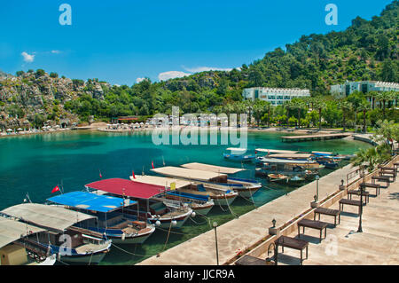 Piccoli taxi turistici barche ormeggiate in fila alla passeggiata lungomare con la spiaggia e gli hotel a sfondo nella piccola cittadina di turunc vicino a Marmaris, Turchia Foto Stock