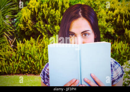 Close up ritratto di donna nascosto dietro libro contro piante e alberi che crescono Foto Stock