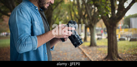 Sorridente fotografo maschio utilizzando telecamere contro il sentiero tra gli alberi in posizione di parcheggio Foto Stock