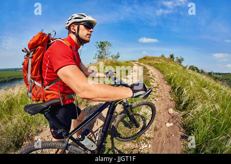 Giovani viaggiatori in maglia rossa con zaino arancione in sella ad una mountain bike su strada di campagna contro il cielo blu Foto Stock