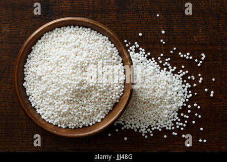 Asciugare le perle di tapioca in legno scuro ciotola isolato in legno marrone scuro dal di sopra. Eventuali fuoriuscite di tapioca. Foto Stock