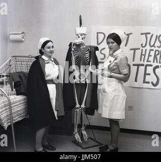 "È ancora vivo?" degli anni sessanta, Inghilterra, su un ospedale, due infermieri femmina con una dimensione di vita scheletro modello vestito con un infermiere del promontorio e hat e un panino nella sua bocca. Una delle infermiere, che pretende di controllare l'impulso dello scheletro in una protesta per sopravvivere sui bassi salari. Foto Stock