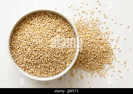 La quinoa semi in ceramica bianca recipiente isolato sul dipinto di bianco legno proveniente da sopra. Eventuali fuoriuscite di semi. Foto Stock