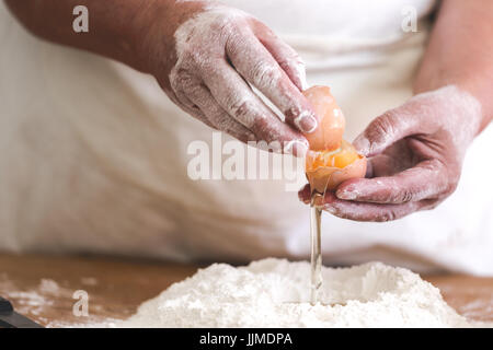 Senior donna aggiunta di uova per la pasticceria. Concentrarsi sulle uova rotte e vecchie mani di lavoro. Orientamento orizzontale, saturi colori sbiaditi Foto Stock