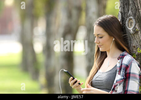 Rilassati ascoltando la ragazza sulla linea di musica e la visione di contenuti multimediali appoggiato su di un albero in un parco Foto Stock