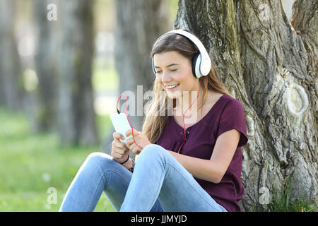 Single teen ascoltare musica on line con le cuffie e smart phone seduta sul prato di un parco Foto Stock