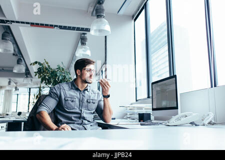 Grave giovane imprenditore seduto e parlando al telefono cellulare in ufficio. Professionisti creativi seduto al suo posto di lavoro. Foto Stock