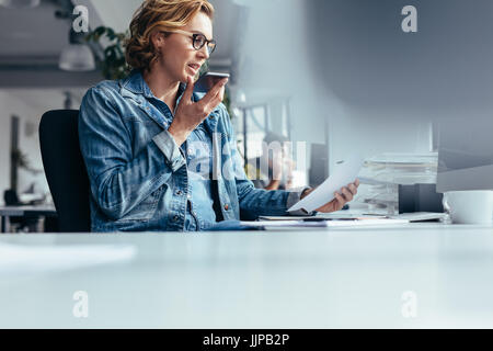 Giovane imprenditrice parlando al telefono con altoparlante e documento. Giovane donna seduta in ufficio con smart phone e guardando una carta. Foto Stock