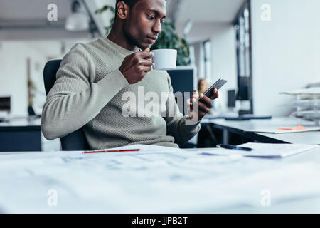 Imprenditore seduto alla scrivania in ufficio un caffè. Giovane africano con la tazza di caffè e smart phone. Foto Stock