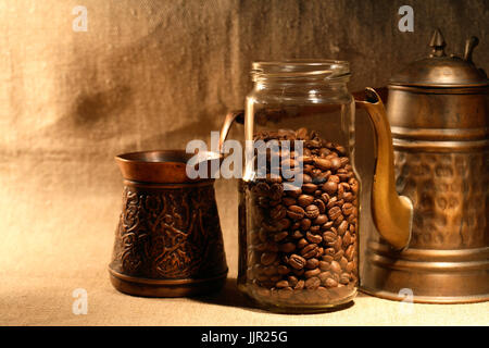Vaso di vetro con i chicchi di caffè nei pressi della vecchia caffettiera in rame su tela lo sfondo Foto Stock