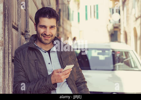 Uomo sorridente in possesso di un telefono cellulare all'aperto in piedi accanto alla sua nuova vettura texting. Foto Stock