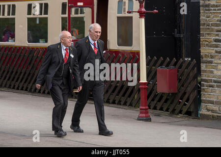 Due maschi guardie del treno in uniforme, camminando insieme lungo la piattaforma - storica stazione Oxenhope, Keighley & Worth Valley Railway Station, Inghilterra, Regno Unito. Foto Stock