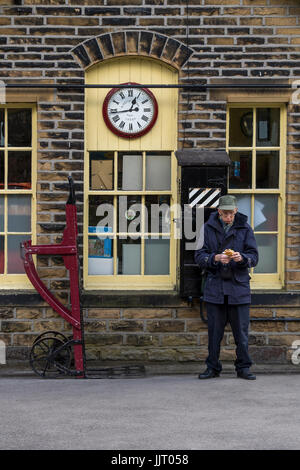 L'uomo accanto al vecchio ufficio booking, orologio e bagagli carrello sulla piattaforma - storica stazione Oxenhope, Keighley & Worth Valley Railway Station, Inghilterra, Regno Unito. Foto Stock