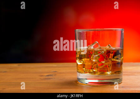 Un bicchiere di whisky con ghiaccio su un tavolo di legno contro un nero e sfondo rosso con spazio di copia Foto Stock