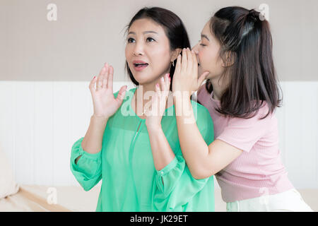 Asian ragazza adolescente si sussurra a sua madre orecchio mentre sono di trascorrere del tempo insieme a casa. Madre cercando sorpreso. Foto Stock