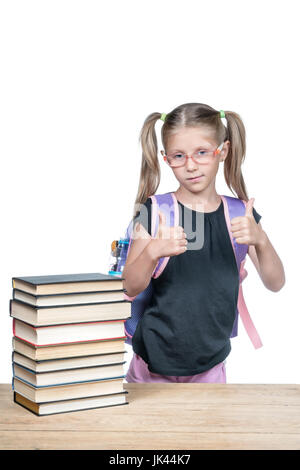 Schoolgirl in bicchieri con zaino mostra due mani Thumbs up, sta in piedi in una scrivania di legno con una pila di libri, isolati su sfondo bianco Foto Stock