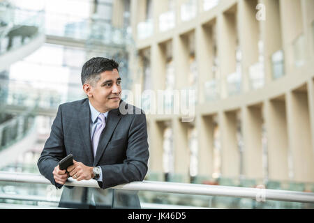 Razza mista imprenditore appoggiata sulla ringhiera in lobby azienda telefono cellulare Foto Stock
