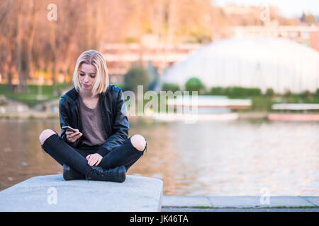 Caucasian donna seduta sulla parete vicino al fiume sms sul cellulare Foto Stock