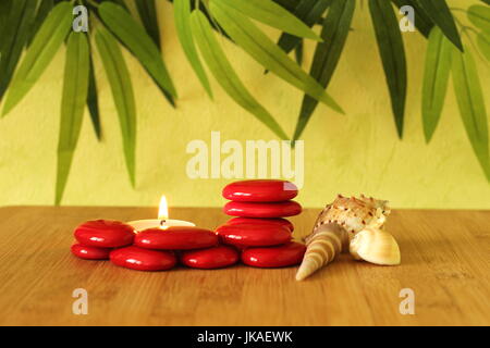 Piccole pietre rosse disposte in colonne in stile zen con una candela e conchiglie sulla spiaggia di bambù sul pavimento in legno e il fogliame verde sullo sfondo Foto Stock