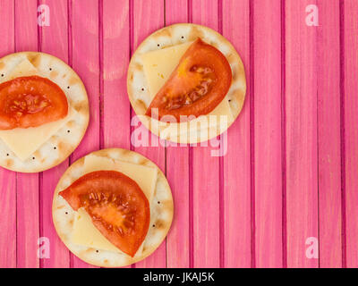 Formaggio e pomodoro su biscotto acqua crackers contro una rosa di sfondo di legno Foto Stock