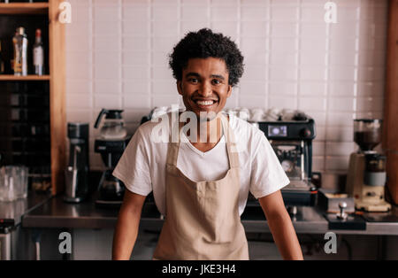 Ritratto di felice giovane barista permanente al cafe contatore. L'uomo africano nel grembiule guardando la fotocamera e sorridente. Foto Stock