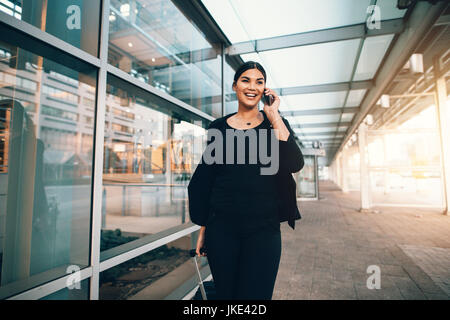 Felice giovane donna in viaggio d'affari a piedi con i bagagli e parlando al cellulare all'aeroporto. Viaggiare imprenditrice effettuare chiamate telefoniche.