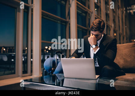 Stanco imprenditore in attesa del volo ritardato nella sala di attesa di un aeroporto. Giovane uomo seduto in sala d'attesa con laptop sensazione di esaurimento. Foto Stock