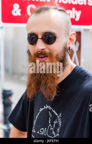 Ritratto, viso uomo caucasico, 20s, marrone lunga barba e capelli corti, tondo nero occhiali da sole. Prigioniero di metallo piercing mento e naso. Contatto visivo. Foto Stock