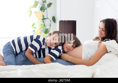 Giovane bella donna incinta e il suo dolce bambini, twin boys, cuddling con amore e tenerezza, seduta sul letto in camera da letto, attesa con impazienza fo Foto Stock