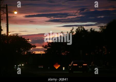 Guida auto lungo la strada di quartiere dopo il tramonto sotto rosso grigio e blu nuvole, le sue luci di illuminazione profilo a rombo arancione lavori stradali Segno Foto Stock