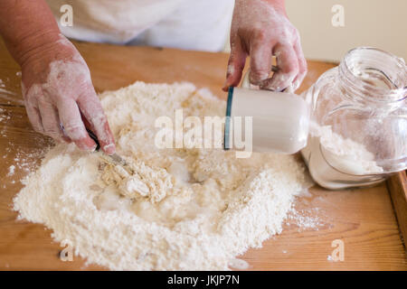 Senior donna rende la pasta con la farina e versare dell'acqua. Vista dall'alto, il fuoco selettivo sulla mano di lavoro con il coltello. Raccolto orizzontale Foto Stock